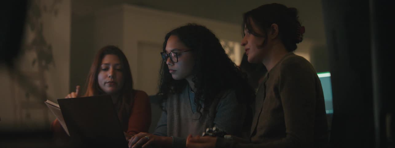 Three women collaborating on a laptop in a dimly lit room engaged in focused discussion