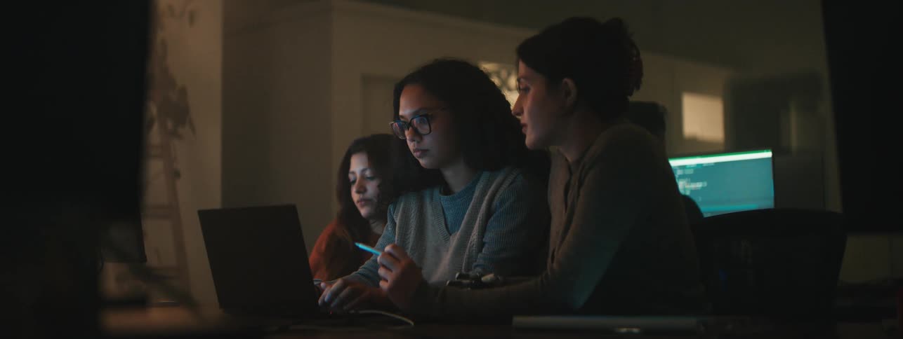 Group of three women sitting in front of a laptop in a lowlit room