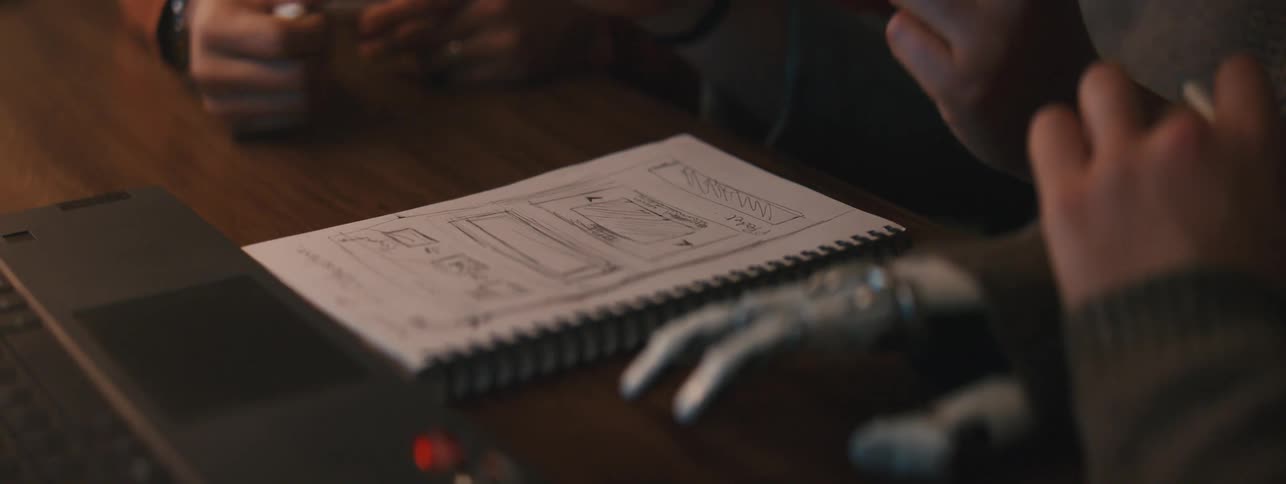 Three people at a table and gesture over drawings in a small notebook