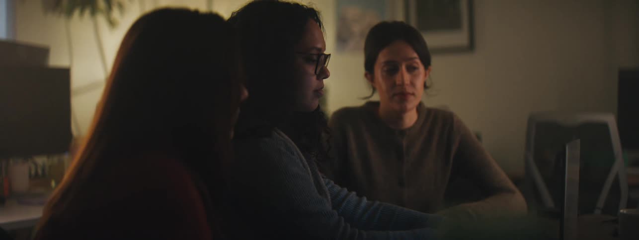 Three women engaged in a discussion in a dimly lit office setting