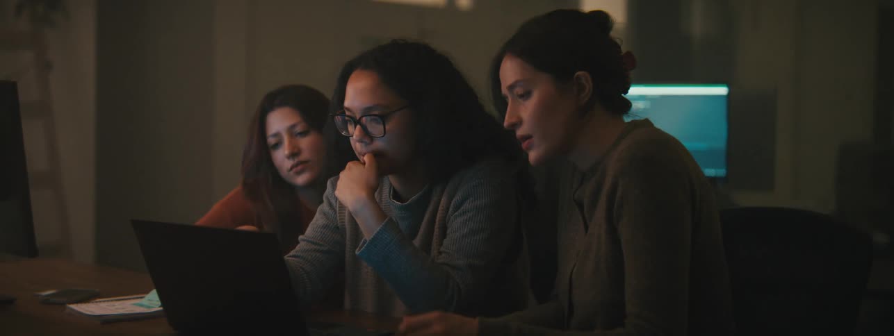 Three women collaborating intently around a laptop in a dimly lit workspace