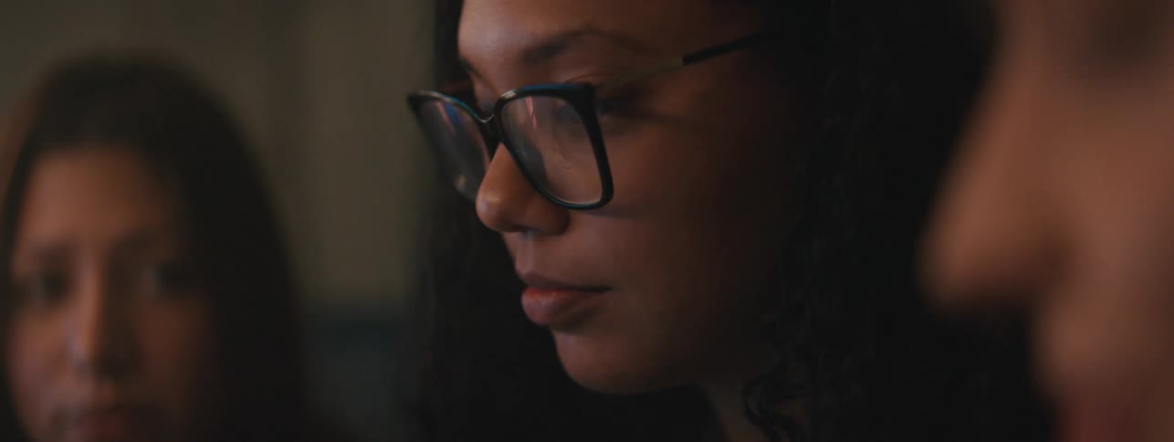 Portrait of a woman wearing glasses sitting at a table next to two other women in a lowlight environment
