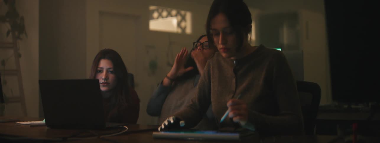 A group of students sitting at a table with laptops and tablets in a dimly lit room working late