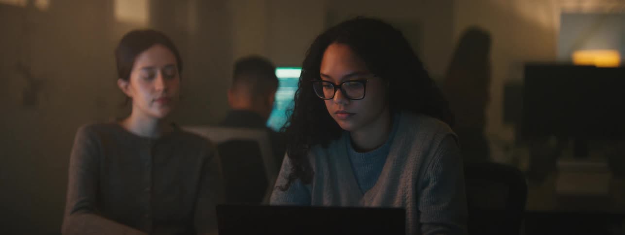 Two women sitting at a desk talking while focused on a laptop with a man sitting behind them at a computer