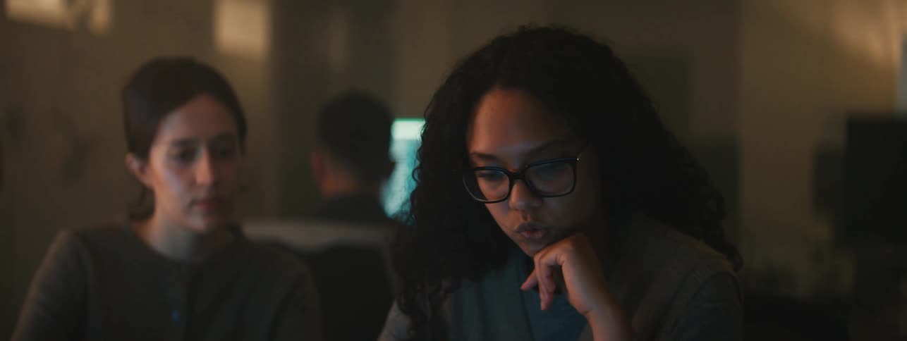Two women focused on a computer screen in a dimly lit environment having a discussion
