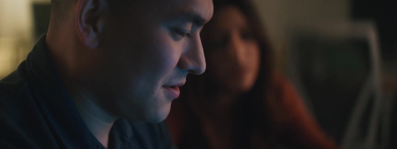 A man and a woman smiling nodding and talking while looking at a computer that lights up their faces