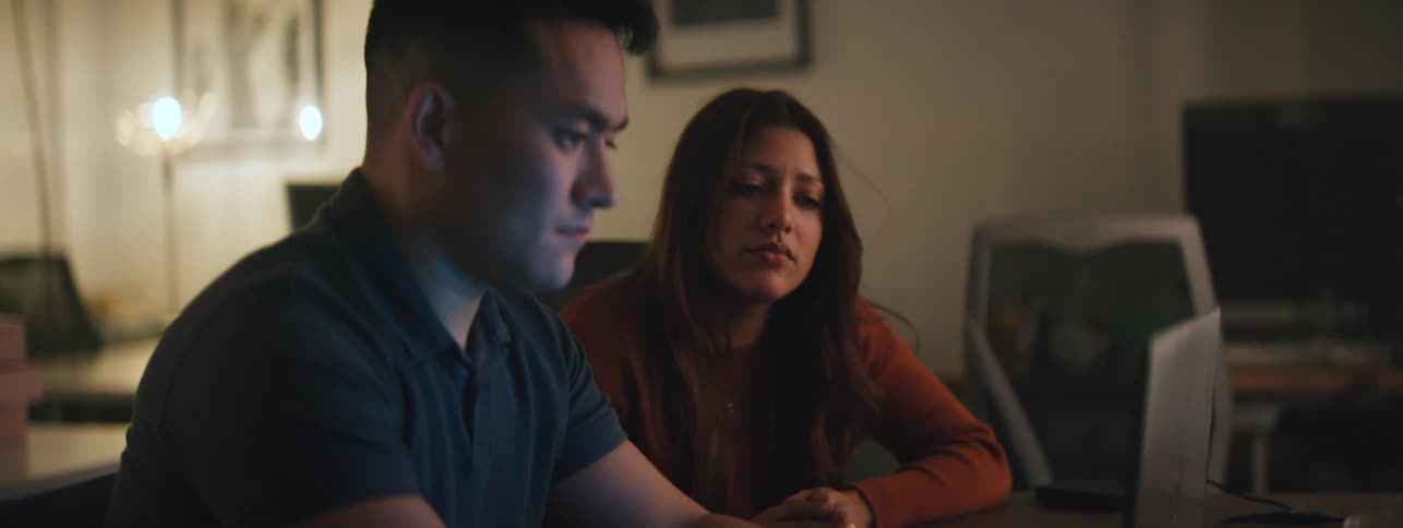 A woman and a man talking while looking at a laptop then he runs his hands through his hair while sighing before turning back to the laptop and typing