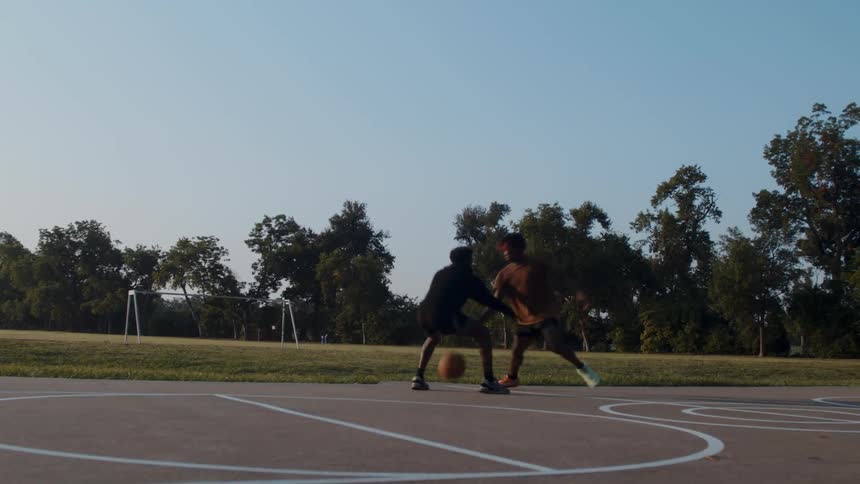 Two black men are playing basketball on an outdoor court on a summer day