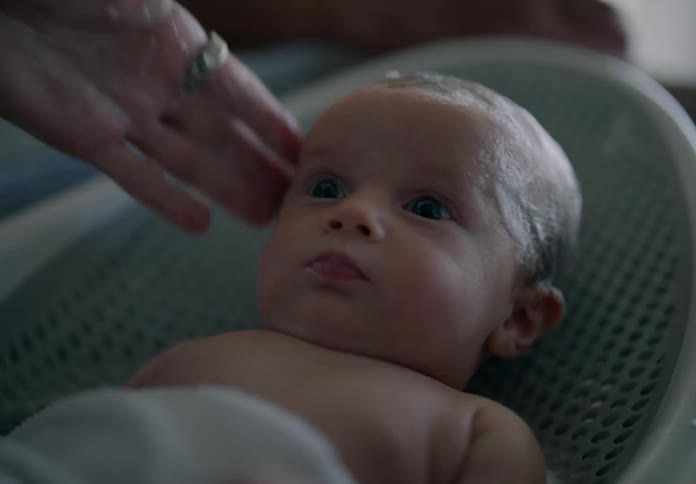 A baby lying down has shampoo on his head and next to him is an adult hand that is bathing him