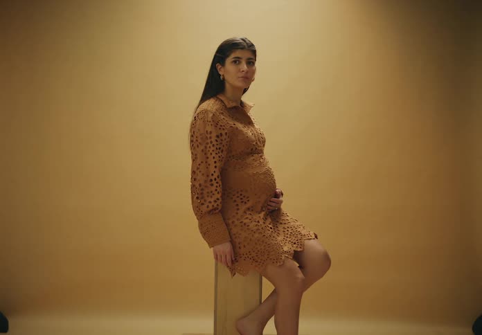 A woman in a brown dress is pregnant and sitting with her hand on her belly and is in an environment with an orange background