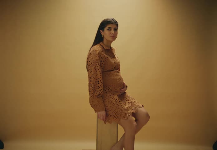 A pregnant woman seated on a wooden stool against a beige background wearing a dress