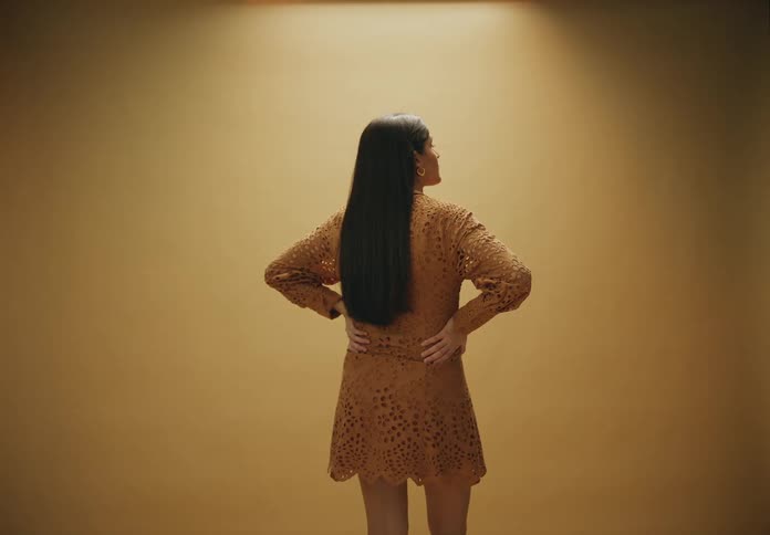 Woman standing seen from behind with black hair and hands on hips wearing a caramel dress