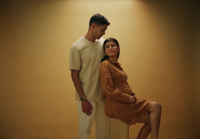 A pregnant couple stand together for a portrait against a beige background