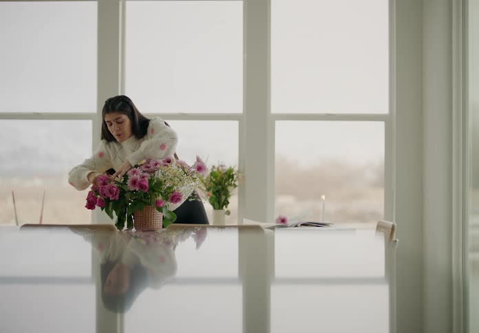 A woman stands next to large windows arranging a bouquet of pink and white flowers
