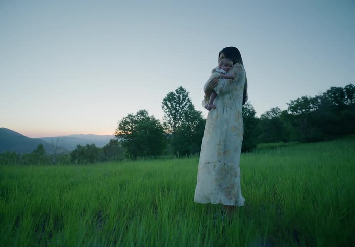 Mother carries her baby through a meadow at sunset