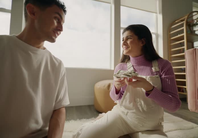 A man and a pregnant woman are sitting on the floor and looking at a pair of baby shoes and they are happy and give each other a little kiss