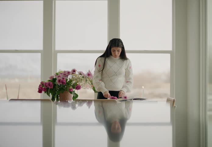 A woman stands next to large windows arranging a bouquet of pink and white flowers