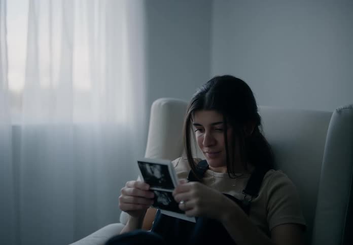 A pregnant woman sitting in a chair by a window with sheer curtains looking at echo sonograms