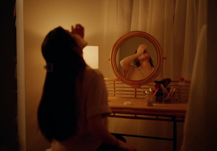 A woman opening a bottle of medicine and taking and then drinking some liquid from a glass while she is looking in the mirror in a dark room with an orange light