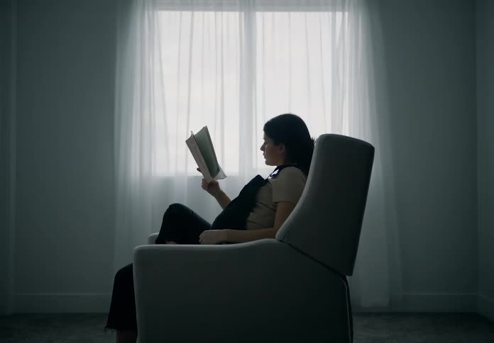 A pregnant woman sitting in an armchair engrossed in reading a book next to a window