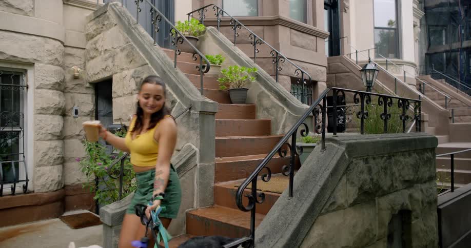 A woman coming down an outside staircase with several dogs on leashes and she is drinking a brown liquid from a glass with a straw