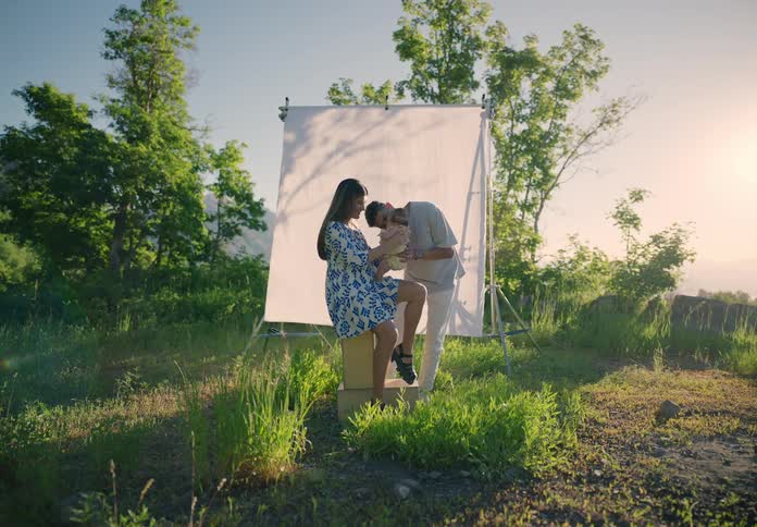 Mother sitting with a baby on her lap and the father next to her while standing on a hill with a white screen in the background