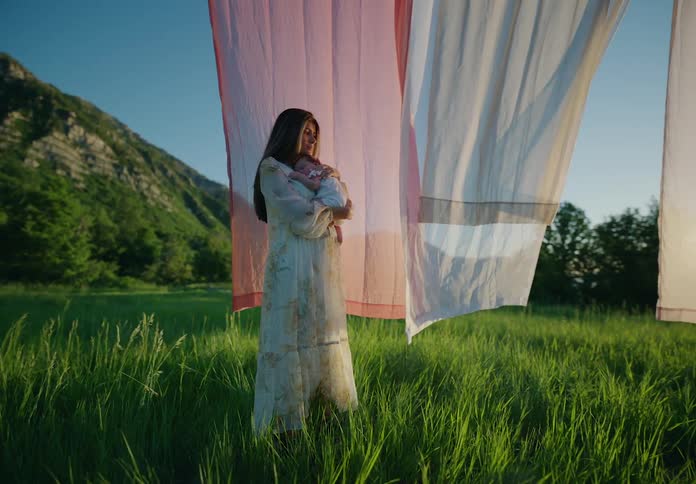 A mother standing in a grassy field holding a baby with clothes hung on a line