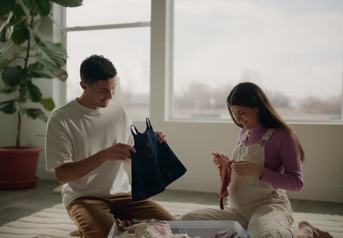 A man and a woman sit on the floor near a window and are happy as they look at baby clothes
