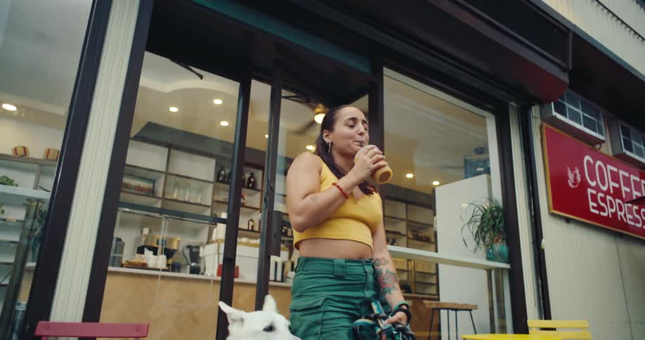 A woman stands outside a coffee shop holding a cup and walking dogs in the city
