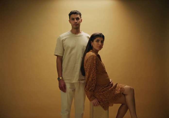 A pregnant woman seated on a wooden stool against a beige background a man standing besides her