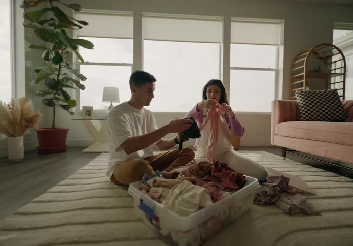 A couple sitting on the floor folding laundry from a basket in a welllit cozy living room with plants and furniture