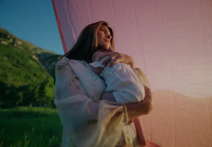 Mother lovingly holding a newborn baby in her lap while standing in a natural setting with a blanket in the background