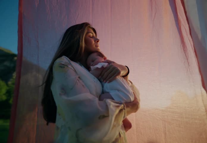 Mother stands in a field near the mountains swaying with her baby as nearby hanging sheets flutter in the breeze