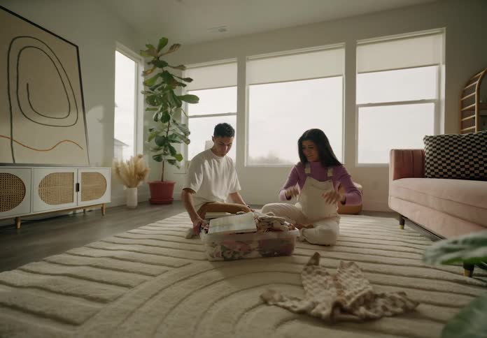 A man placing a basin of baby clothes on the floor where there is a woman sitting and she happily picks up the clothes and throws them forward