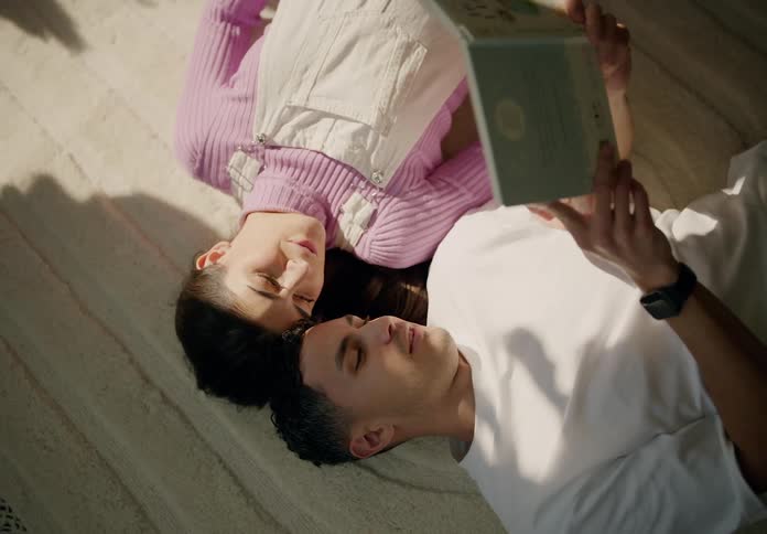 A couple lies on the bed their heads resting against each other reading a book in the morning