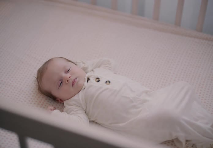 A little baby is almost asleep and he is lying in a crib with a white sheet underneath
