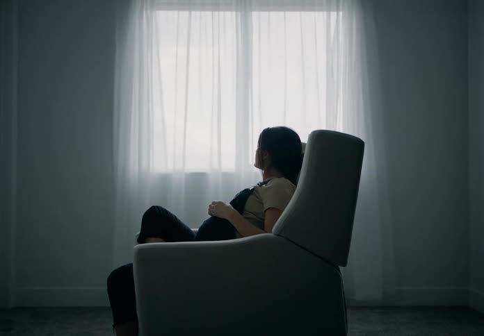 A pregnant woman sitting in a chair by a window with sheer curtains