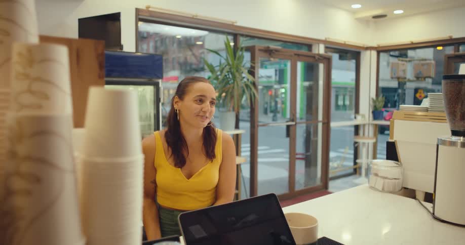 A woman in a yellow top engaged in conversation at a cafe while a barista is partially visible in the foreground
