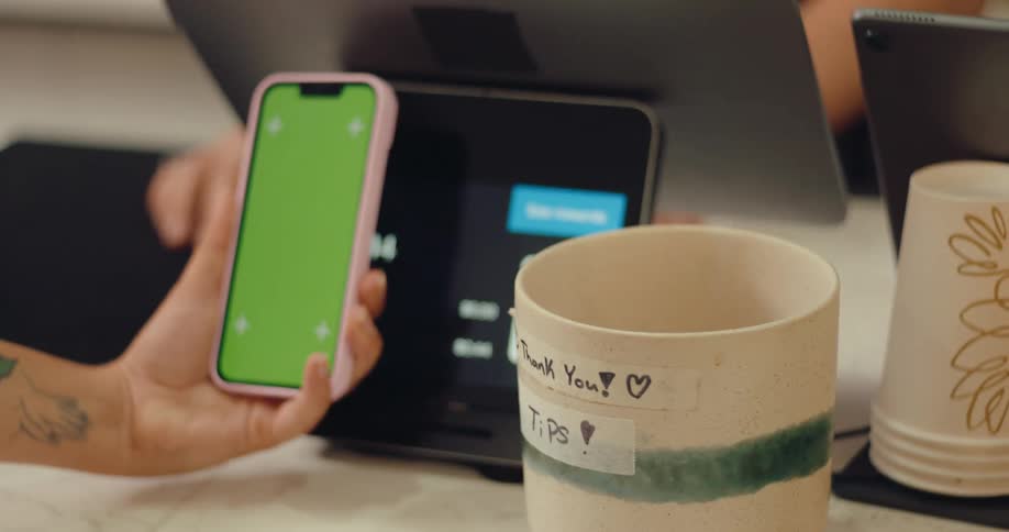 A hand holds a smartphone with a green screen in front of a payment processor