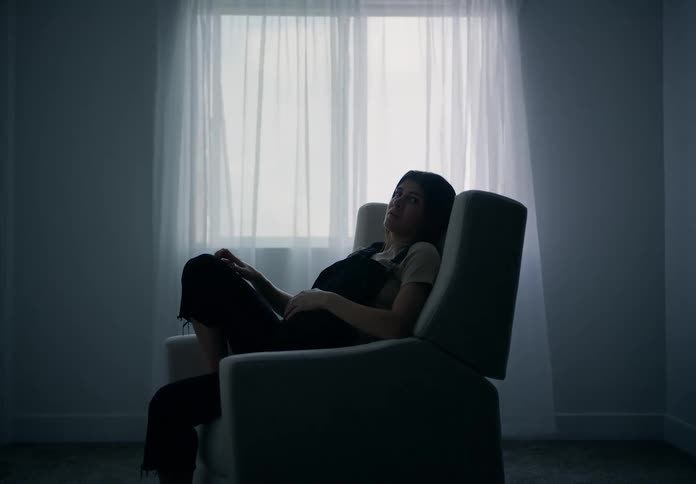 A woman sitting in an armchair next to a window with a white curtain was pointing forward as a person passes by and she is pregnant