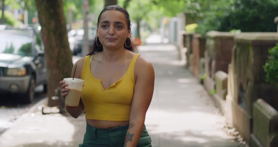Woman sips a coffee drink while walking a dog along a residential sidewalk
