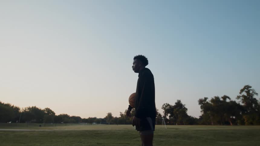 A young man stands on a park at dusk with a basketball under his arm