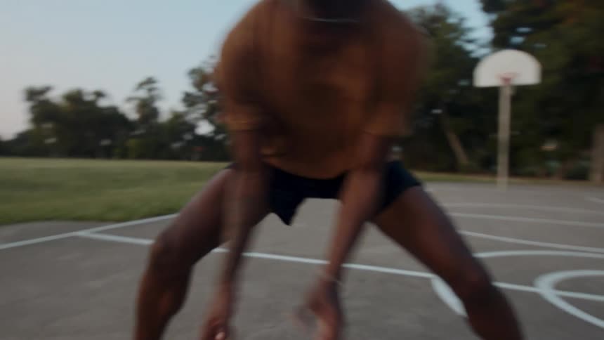 A young man dribbling a basketball on an outdoor court during early evening