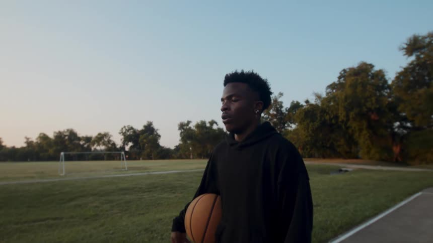 A young man stands still holding a basketball on a court at sunset