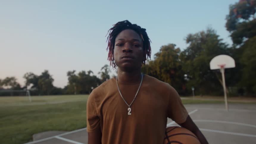 A young man stands on a court in the park with a basketball under his arm