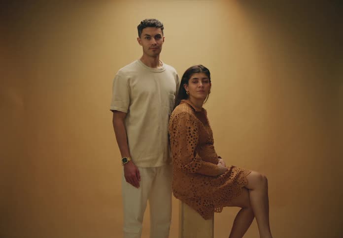 Man and woman model clothing against a partially lit brown wall