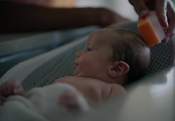 Baby lying in a bathtub in the sink with a persons hand holding a bar of soap and running it through his hair