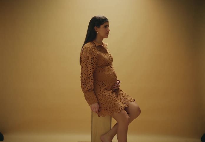 A pregnant woman seated on a wooden stool against a beige background wearing a dress