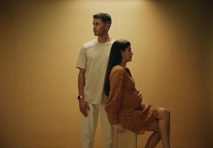 A pregnant woman seated on a wooden stool with a man standing next to her against a beige background wearing a dress