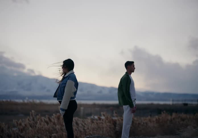 Man and a pregnant woman stand back to back in a field with mountains in the background
